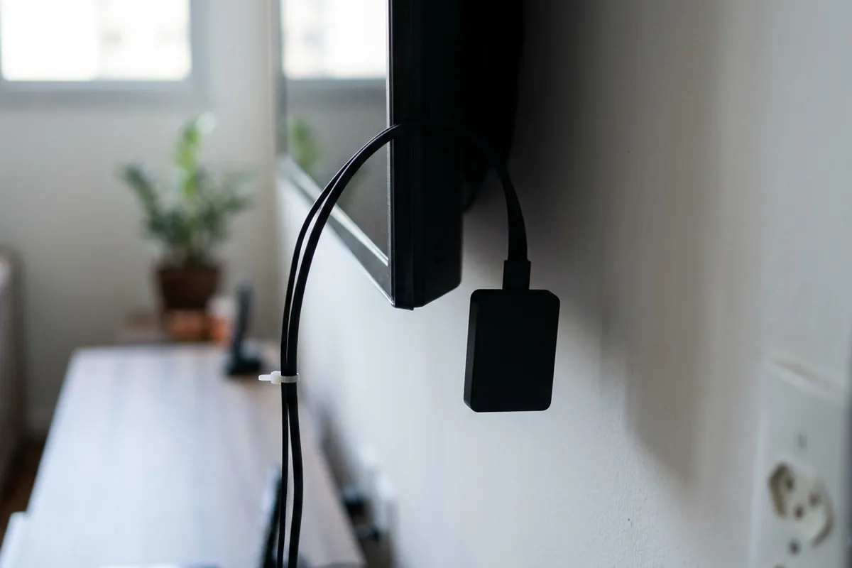 Dongle preto pendurado num cabo curto que sai de trás de uma TV na parede, com os cabos presos por abraçadeira