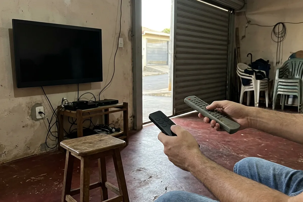 Duas mãos segurando controles diferentes diante de uma TV apagada numa garagem convertida
